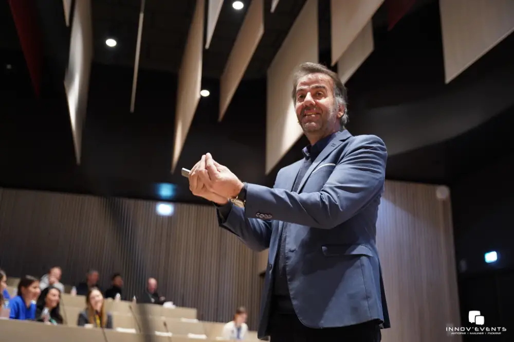 Homme en costume parlant avec enthousiasme devant une assemblée lors d'une conférence à Pau, dans une salle moderne avec plafond acoustique suspendu.