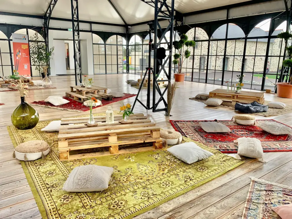 Aménagement d’un espace événementiel zen avec des tapis colorés, coussins au sol et tables basses en palettes dans un espace verrière lumineux