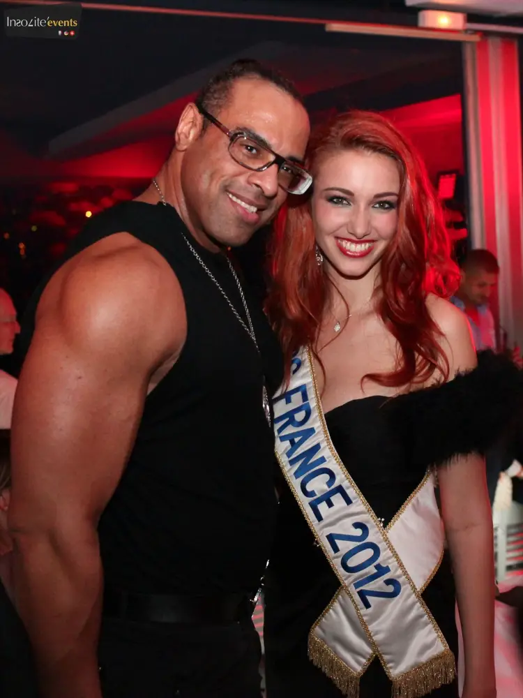 Miss France 2012 souriante aux côtés d'un invité musclé lors d’une soirée événementielle mode dans une ambiance rouge chic