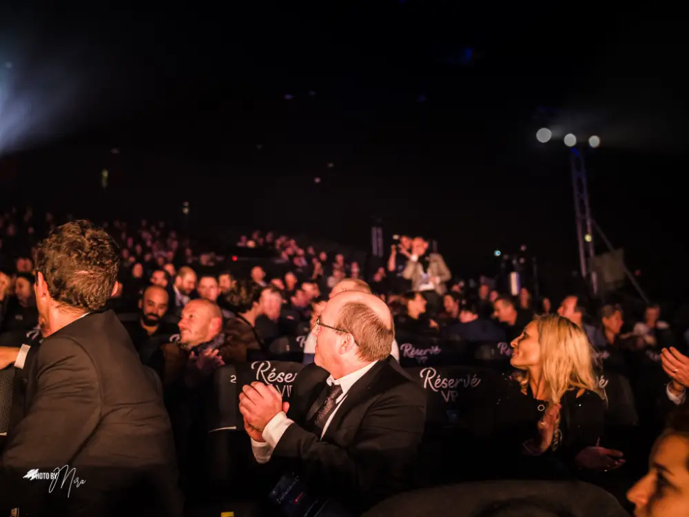 Un homme en costume applaudit debout dans une salle de conférence comble, ambiance tamisée et festive.