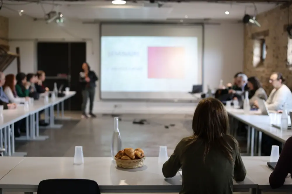 Participants assis en U autour de tables blanches lors d’un atelier ou séminaire avec projection au fond.