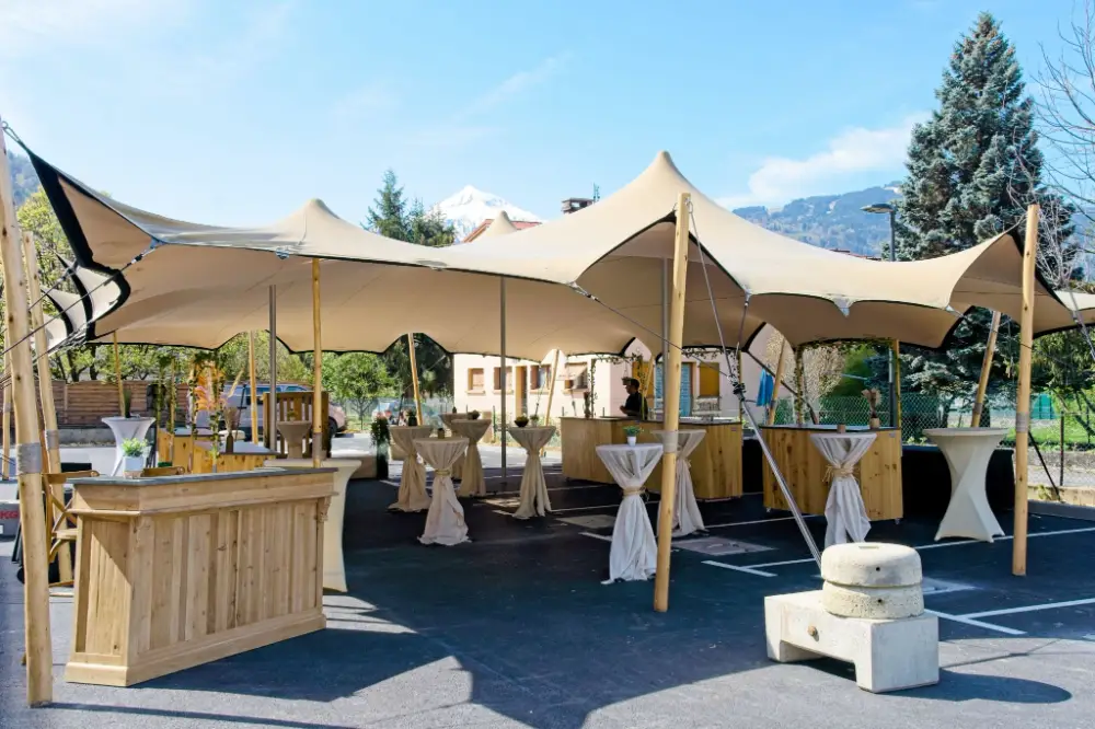 Scénographie extérieure avec tente stretch, mange-debout drapés et bars en bois pour un événement d’entreprise en plein air avec vue sur les montagnes