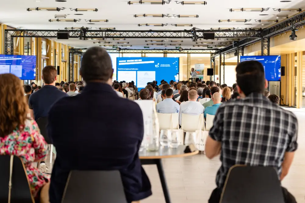 Vue arrière d’un public attentif pendant une présentation professionnelle dans un espace lumineux et moderne.